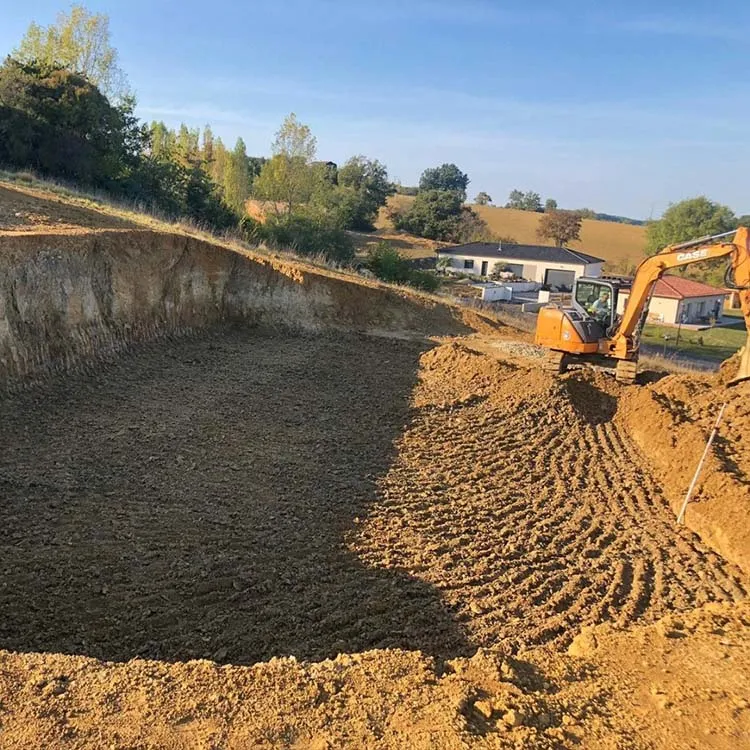 Pelle mécanique en pleine opération de terrassement sur un chantier en pente, avec des bâtiments en arrière-plan. ​​